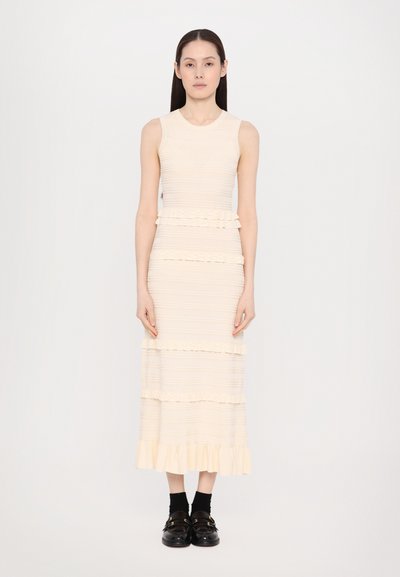 Light beige sleeveless dress with textured stripes and ruffled tiers at the waist and hem, paired with black shoes and ankle socks.