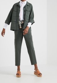 Groene utility jack met knoopsluiting en meerdere zakken, gecombineerd met bijpassende broek. Bruine platform sandalen. Witte gestreepte shirt.