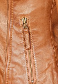 Tan leather jacket section featuring a zipper enclosure with gold-toned hardware, visible stitching, and a textured surface.