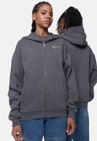 Grå zip-hoodie lavet af blødt stof, med en kængurulomme, justerbar hætte og broderet logo på venstre bryst.