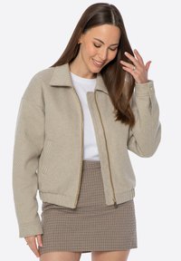 Giacca bomber beige a righe con chiusura frontale a zip, ampio colletto e maniche con polsini, abbinata a una minigonna a quadri.