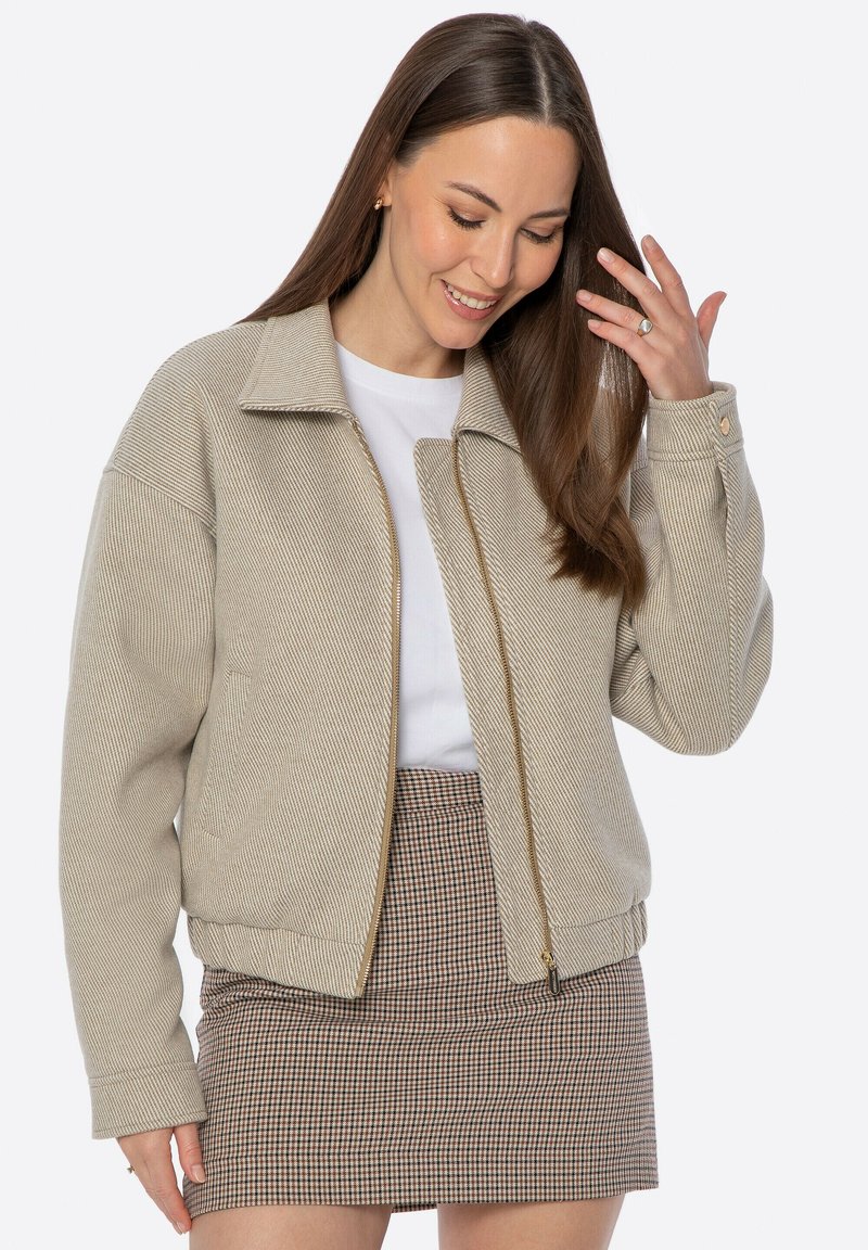 Giacca bomber beige a righe con chiusura frontale a zip, ampio colletto e maniche con polsini, abbinata a una minigonna a quadri.