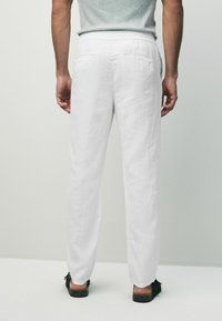 White linen trousers with a relaxed fit, featuring a standard waistband and two back pockets. Worn with black slip-on shoes.