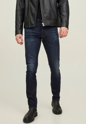 Mann trägt dunkelblaue Slim-Fit-Jeans, schwarze Lederjacke und schwarze Knöchelstiefel, steht vor einem einfarbigen Hintergrund.