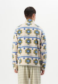 Mies, joka on pukeutunut vaaleanbeigeen fleece-takkia, jossa on sinisiä ja vihreitä geometrisia kuvioita, yhdistettynä vaaleanvihreisiin ruutuhousuihin, kääntyy poispäin valkoista taustaa vasten.