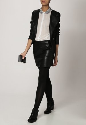 Kvinde iført en sort læderjakke og nederdel, hvid bluse med mønstret krave, sorte strømpebukser, ankellange støvler, holder en sort og hvid clutch.