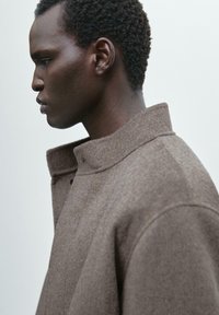 Brown woolen coat with a high collar and subtle texture, featuring a relaxed fit and minimalistic design, shown in profile against a light background.