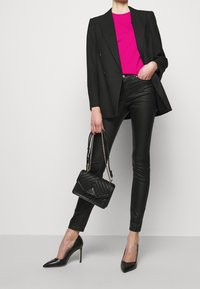 Mujer con blazer negro, top rosa, pantalones de cuero negros, sosteniendo un bolso negro acolchado, usando tacones altos negros, de pie contra un fondo blanco.