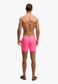 Homme debout, dos au spectateur, portant un short de bain rose vif et des sandales marron sur un fond blanc uni.