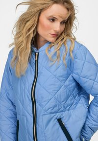 Svetle modrá prošívaná bunda má kapuci a kontrastní námořnický zip. Tkanina má hladkou strukturu s diagonálním prošíváním a bočními kapsami.