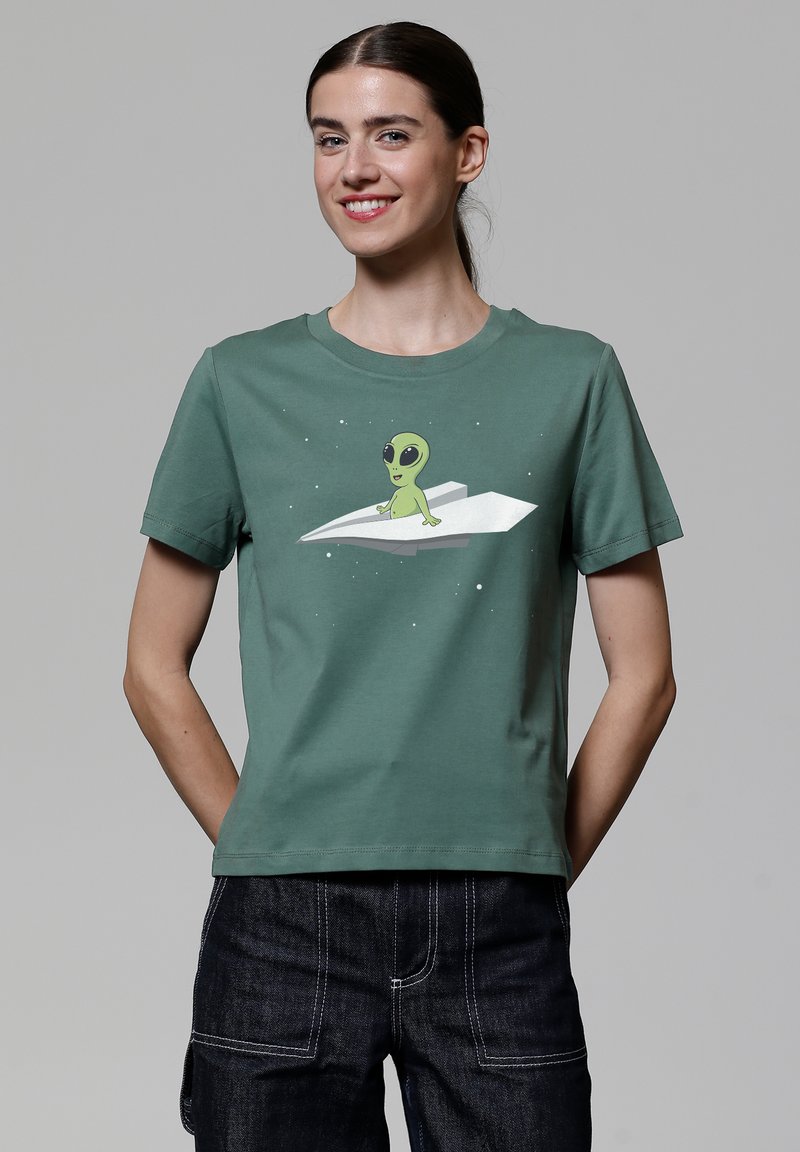 Grünes Baumwoll-T-Shirt mit einem Cartoon-Alien auf einem Papierflugzeug, vor einem sternenklaren Hintergrund. Kurze Ärmel, runder Halsauschnitt.