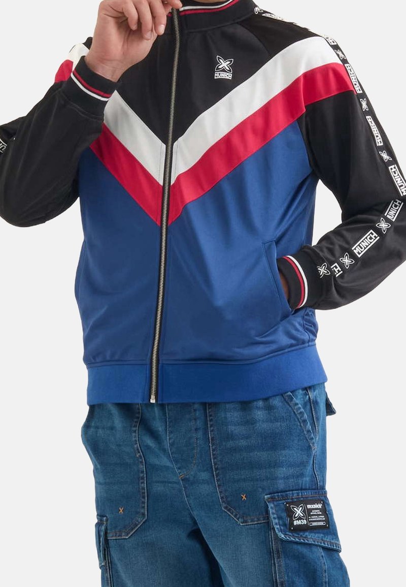 Homme portant une veste zippée bleu, noir, rouge et blanc avec des manches marquées et un pantalon cargo en denim bleu avec une pièce de poche latérale.