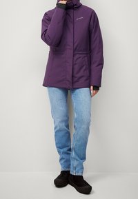 Fioletowa kurtka ocieplana z wysokim kołnierzem, elastycznym pasem i dwoma bocznymi kieszeniami, zestawiona z jasnoniebieskimi dżinsami i czarnym obuwiem.