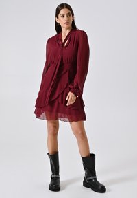 Bordeauxfarbenes Kleid mit langen Ärmeln, gebundenem Ausschnitt und geschichtetem Rüschensaum. Kombiniert mit schwarzen kniehohen Stiefeln mit metallischen Akzenten.