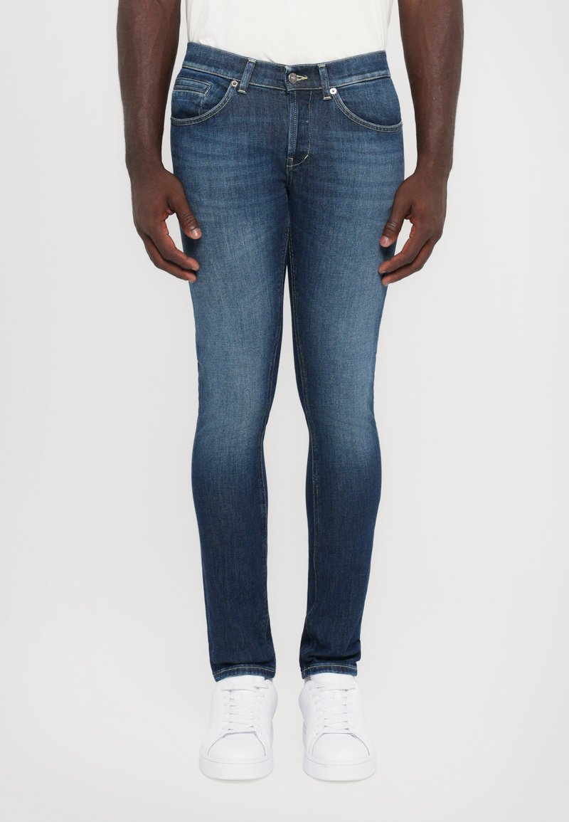 Man som bär slim-fit blå jeans och vita sneakers, stående mot en enkel ljus bakgrund med avslappnade händer vid sidorna.