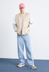 Uomo con cappellino rosa, gilet beige con zip, felpa grigia chiara, pantaloni azzurri e sneakers beige che si trova su un tappeto blu.