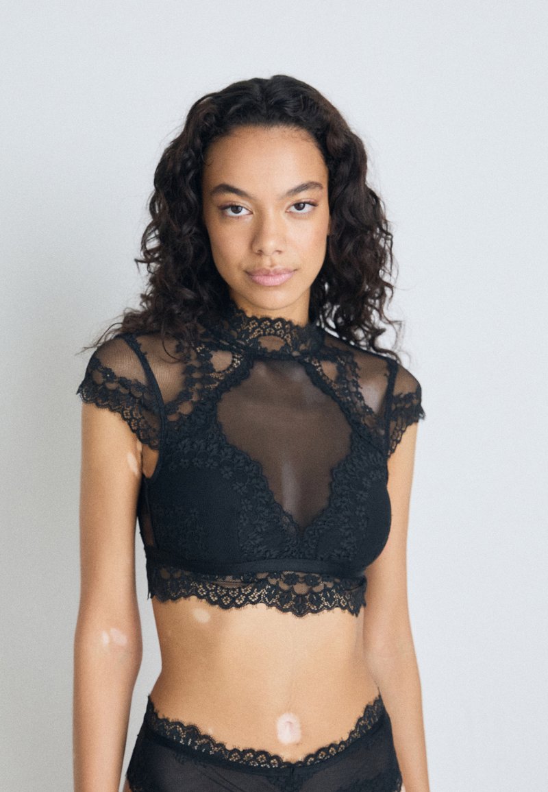 Svart blondebralette med gjennomsiktige mesh-paneler, med høy hals og bølgede kanter. Mykt, teksturert stoff med blomsterdesign.