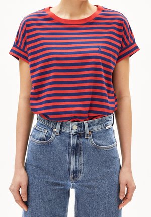 Nainen pukeutuneena punaisen ja tummansinisen vaakaraidalliseen lyhythihaiseen paitaan, joka on tungettu korkeavyötäröisiin sinisiin farkkuihin, seisoo käsivarret rentoina.