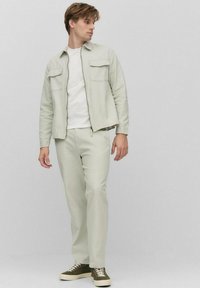 Lichtgroene jas met twee borstzakken, wit shirt en bijpassende lichtgroene broek. Sneakers in donkergroen met beige accenten.