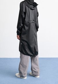 Langer schwarzer wasserdichter Mantel mit Kapuze, tailliert und mit seitlichen Schlitzen. Verfügt über glatte Textur und elastische Bündchen. Kombiniert mit grauen Hosen mit weitem Bein.