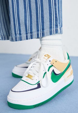 Zapatillas Nike blancas con detalles en verde, panel lateral beige y herrajes dorados. Presentan una suela texturizada y puntera perforada.
