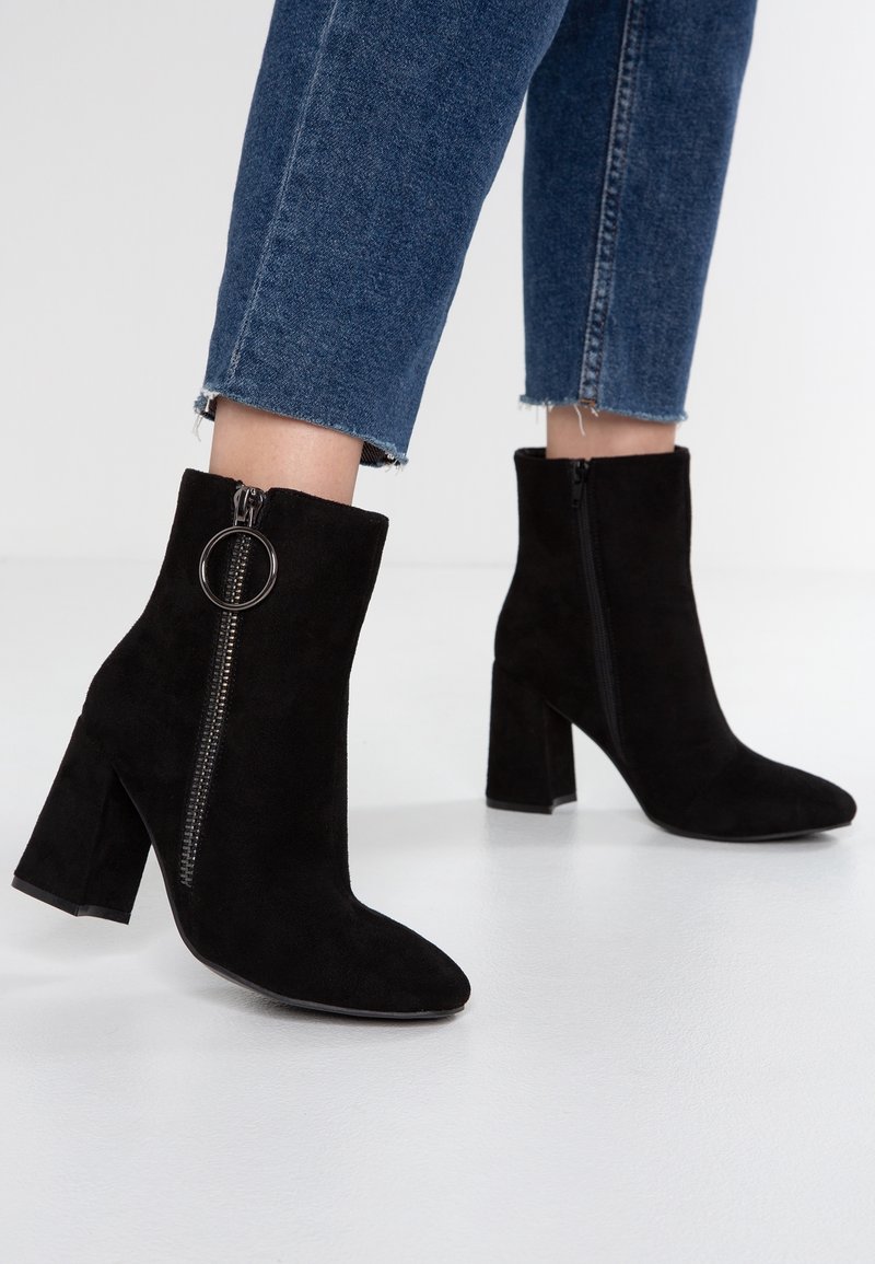 Botas de tobillo de ante negro con puntera puntiaguda, tacón cuadrado y cremallera lateral con un tirador en forma de círculo. Llevadas con jeans azules cortos.