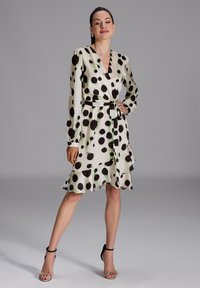 Robe portefeuille en tissu léger, à grands pois noirs sur fond crème, à manches longues et avec un ourlet volanté. Ceinture noire incluse.