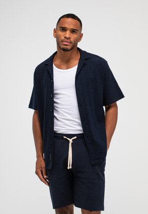 Mann trägt navyfarbenes kurzärmeliges Hemd mit Knöpfen, weißes T-Shirt und navyfarbenen Shorts mit weißem Kordelzug, steht vor einfarbigem Hintergrund.