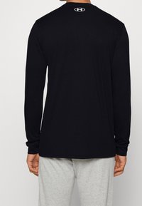 Chemise noire à manches longues en coton, avec un col rond et un logo au dos. Associée à un pantalon gris clair.