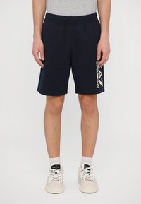 Navy shorts made of jersey fabric with an elastic waistband. Features white logo print along the side. Worn with beige sneakers and socks.