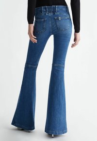 Jeans in denim blu a zampa con vita alta, caratterizzati da due tasche posteriori e una texture liscia. L'orlo termina alla caviglia con una leggera svasatura.