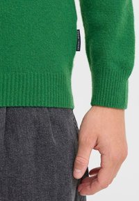 Zielony dziany sweter z prążkowanym mankietem i materiałową metką z napisem "FRIDAY." Towarzyszą mu ciemnoszare spodnie o fakturze.