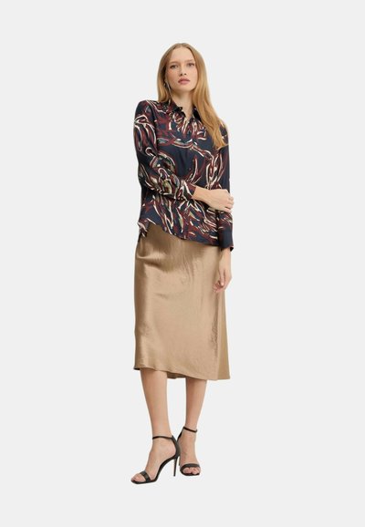 Donna che indossa una blusa a maniche lunghe con motivo astratto, gonna midi di seta color oro e sandali neri con tacco alto, in piedi con le braccia incrociate.