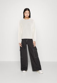 Sudadera crema con un logo texturizado, combinada con pantalones cargo negros de pierna ancha que tienen bolsillos frontales y detalles de costura blanca.