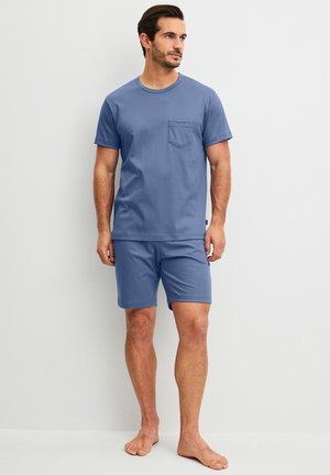 Man staat blootsvoets, draagt een blauw kortemouwhemd met zakje en bijpassende blauwe korte broek tegen een effen achtergrond.