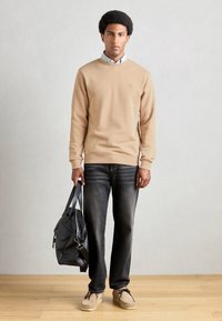 Sweatshirt beige avec des poignets côtelés, jeans foncés et chaussures châtaigne. Tient un sac en cuir noir avec des poches extérieures et une fermeture éclair.