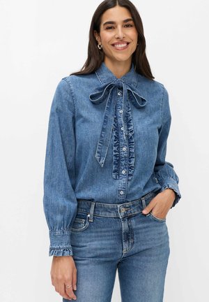 Donna sorridente che indossa una camicetta di denim blu con colletto a fiocco e fronte increspata, abbinata a jeans di denim blu, mano in tasca.