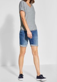 Szara koszulka na krótki rękaw z dekoltem w serek, zestawiona z niebieskimi szortami dżinsowymi z podwiniętym obszyciem, do tego ciemne trampki z białymi podeszwami.