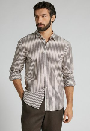 Mand med skæg iført en brun- og hvid-vertikalt striped skjorte med langeærmer og mørke bukser, stående med den ene hånd i lommen.