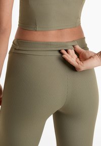 Leggings ribbate de culoare verde măslin cu talie înaltă. Dispun de o talie pliabilă și o textură netedă. Mână plasată la talie.