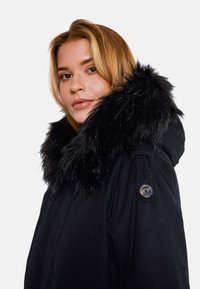 Černá zateplená parka s kapucí z umělé kožešiny. Obsahuje knoflíkový detail na rukávu a hladký matný povrch. Vysoký límec pro teplo.