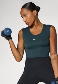 Top sportiv fără mâneci de culoare verde închis cu decolteu rotund și logo-ul Nike, asociat cu colanți negri cu talie înaltă și o ganteră albastră.