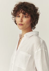 Mujer con cabello corto rizado y marrón, vistiendo una camisa blanca abotonada, mirando con confianza hacia un lado sobre un fondo liso.
