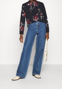 Schwarze Bluse mit floralem Muster und langen Ärmeln, kombiniert mit hochtaillierten, blauen weitem Jeans. Das Model hält eine kleine weiße geflochtene Tasche. Beigefarbene Schuhe.