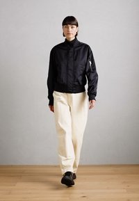 Veste bomber noire avec col haut, poche zippée sur la manche, associée à un pantalon ivoire ample et des chaussures sombres sur un sol en bois.