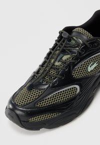 Black and olive green mesh sneaker with lace toggle, thick sole, and green crocodile logo on side, shown from angled top view.