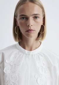 Woman with shoulder-length blonde hair wears large gold hoop earrings and a white buttoned blouse with raised embroidered patterns.