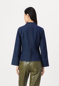 Mörkblå denimjacka med en figursydd design, breda ärmar och kontrasterande sömmar. Bärs över olivgröna byxor i konstläder.