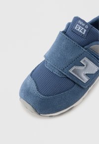 Kék színű velúr sportcipő hálós anyaggal, tépőzárral és fehér gumitalppal. Szürke "N" logóval és 574-es stílusszámmal.