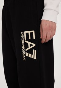 Black sweatpants featuring a large, cream-colored "EA" logo and "EMPORIO ARMANI" text on the side. Soft fabric with an elastic waistband.